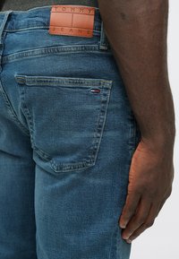 Jeans de ganga azul com um patch de couro a dizer "TOMMY JEANS," apresentando um corte slim e costura visível nos bolsos.