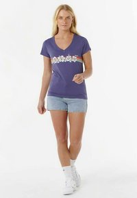 V-neck purple t-shirt with floral design and horizontal stripes, paired with denim shorts and white sneakers, worn by a standing model.