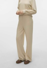 Pantaloni beige a gamba larga realizzati in tessuto morbido, con vita alta e tasche laterali, abbinati a scarpe marroni con stringhe.