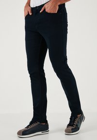 Temno modri, oprijeti kavbojki z gladko teksturo, zasnovo petih žepov in kovinskim gumbom za zapiranje. V kombinaciji s sivimi in mornarsko modrimi športnimi copati.