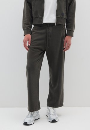 Pantaloni della tuta verde oliva scuro realizzati in misto cotone, con vita elastica, tasche laterali e una vestibilità comoda, abbinati a scarpe da ginnastica bianche.