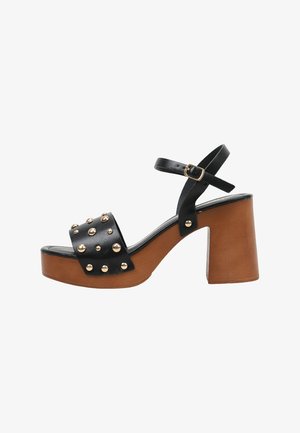 Sandalia de tacón alto de cuero negro con plataforma de madera, tachuelas doradas en la correa delantera y correa ajustable en el tobillo con hebilla dorada.