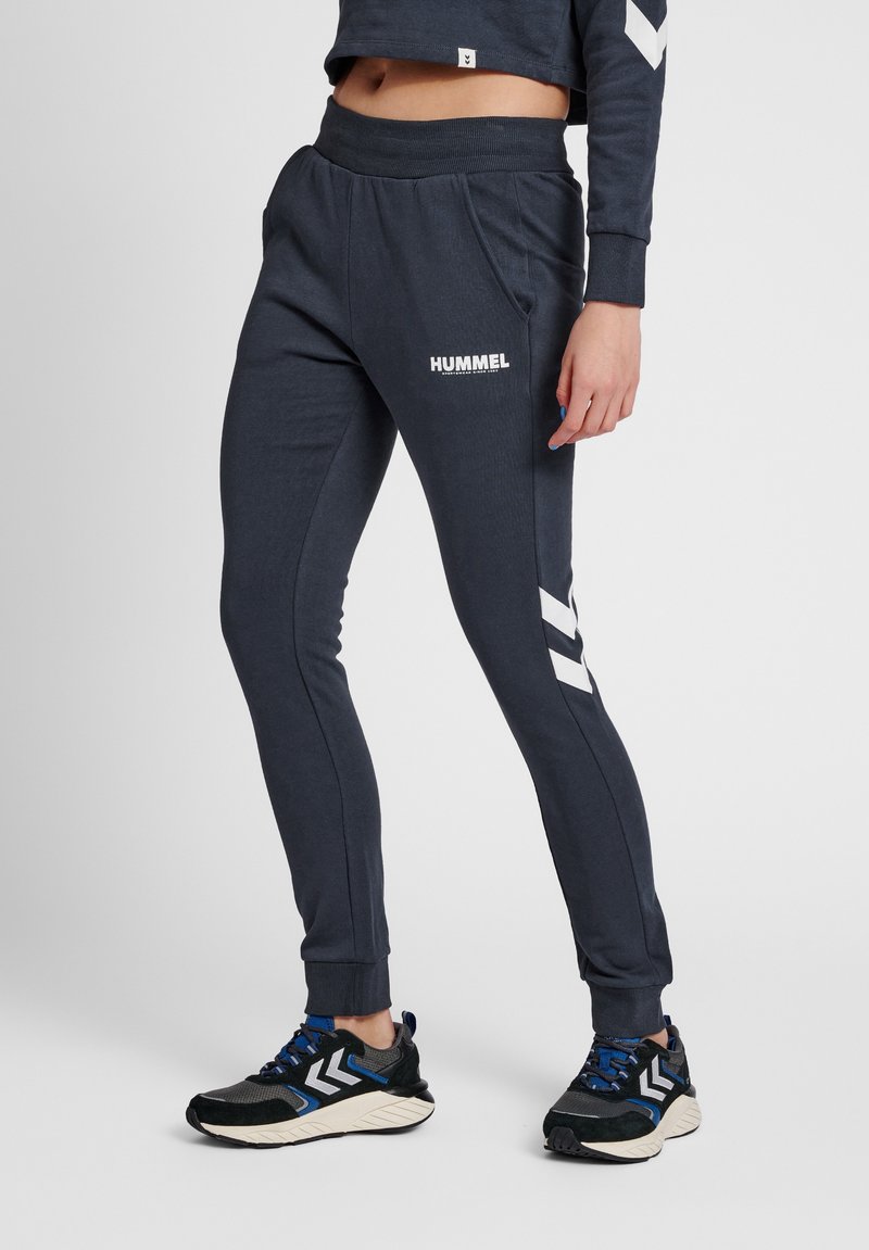 Mörkgrå sweatpants med en åtsittande vrist, prydda med vitt "HUMMEL"-logotyp och chevronmönster. Tillverkade av mjukt material med sidofickor.