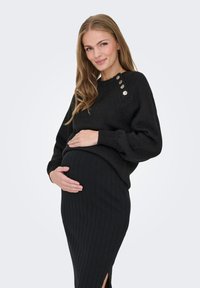 Nosečnica z dolgimi blond lasmi, oblečena v črni pulover z gumbi in črno rebrasto krilo, se smehlja in drži svoj trebuh.