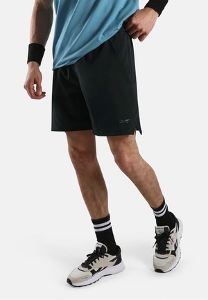 Mann trägt schwarze Sportshorts, blaues Hemd, schwarze Handgelenksbänder, schwarz-weiß gestreifte Socken und beigefarbene sowie schwarze Laufschuhe und steht.