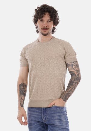 Uomo con capelli ricci che indossa un maglione beige a maniche corte con texture e jeans blu, mostrando tatuaggi dettagliati su entrambe le braccia.
