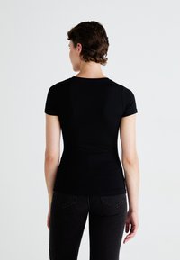 Persoană cu păr scurt, brunet, îmbrăcată într-un tricou negru cu mânecă scurtă și blugi negri, stând cu spatele la un fundal alb simplu.