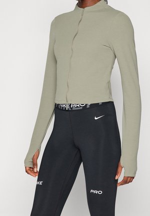 Kobieta ubrana w jasnorozowy, zapinany na zamek, długi top z otworami na kciuki oraz czarne legginsy Nike Pro z logo na talii i nogawkach.