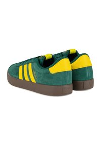 Zelené semišové tenisky se žlutými akcenty a třemi pruhy. Mají hnědou gumovou podrážku a zádový pásek pro snadné obouvání.
