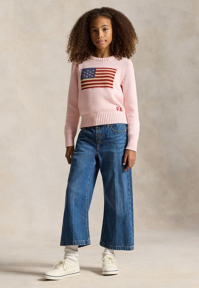 Fille aux cheveux bouclés portant un pull rose avec un drapeau américain, un jean bleu à jambes larges, des chaussettes blanches et des baskets blanches, debout sur un fond neutre.