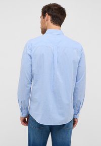 Koszula w niebiesko-białą kratę, z długimi rękawami, mankietami zapinanymi na guziki, prostym dołem i detalami na plecach. Na dole widoczne dżinsy.