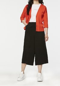 Röd cardigan med trekvartslånga ärmar och knappar, tillsammans med en vit t-shirt med svart text, och svarta vida byxor. Vita sneakers.