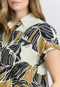 Korte mouwen blouse in lichtgroen met zwart en gouden bladpatroon. Met kraag ontwerp en knoopdetails, gemaakt van lichtgewicht stof.