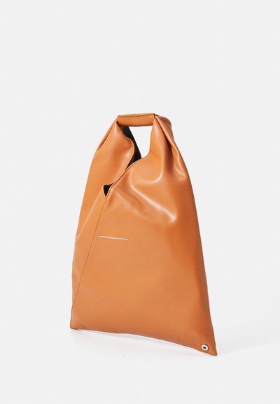 Mjuk tanfärgad läder tote-väska med integrerat handtag och en liten metallöggla i nedre hörnet, stående mot en vit bakgrund.