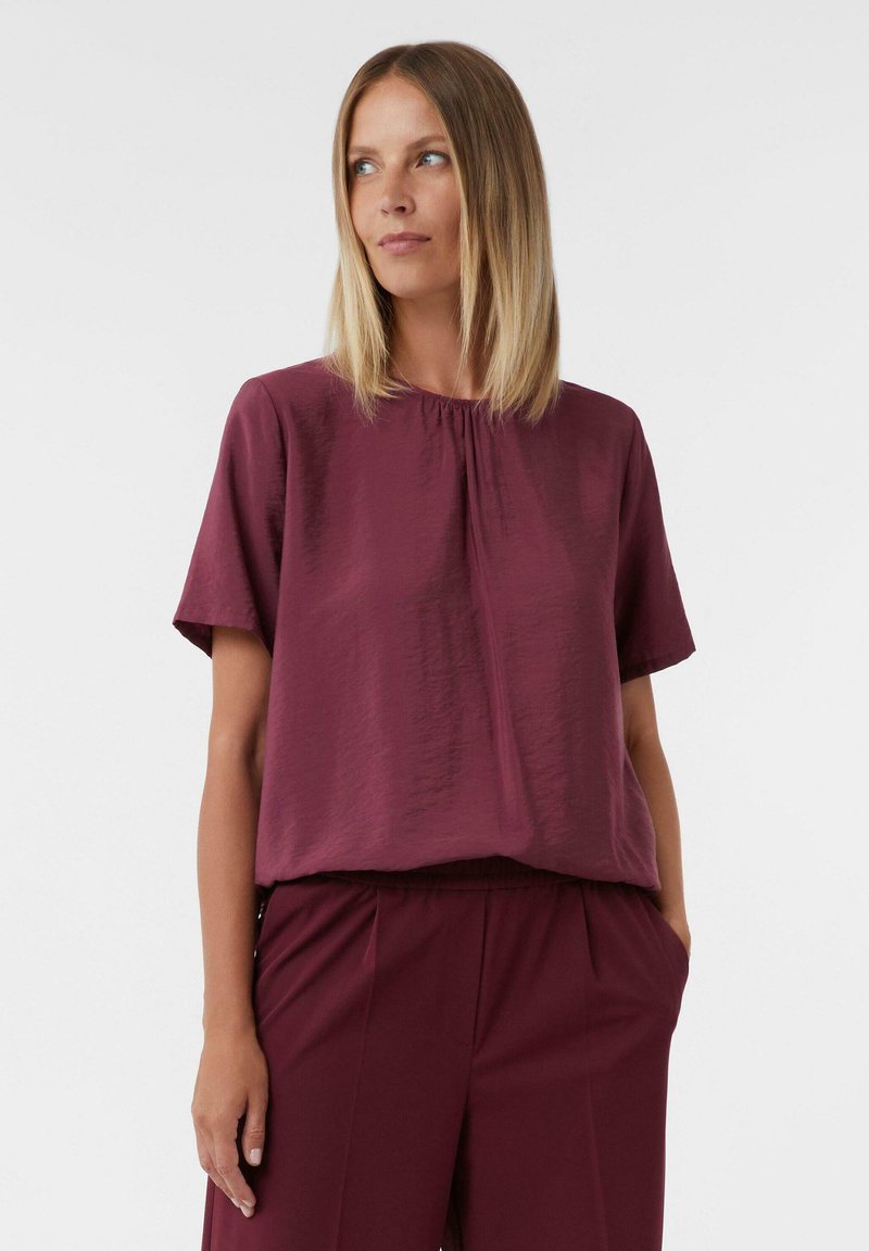 Blusa bordeaux a maniche corte con scollatura arricciata, realizzata in tessuto testurizzato. Indossata con pantaloni a vita alta coordinati, dotati di tasche laterali.