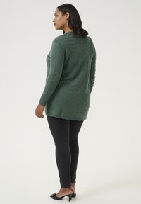 Groene gebreide trui met een zigzagpatroon, lange mouwen en een langere achterkant, gecombineerd met zwarte strakke broek en hakken.
