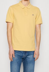 Gul bomullspolo med krage, korta ärmar och en liten marinblå logotyp på vänster bröst, i kombination med blå jeans.