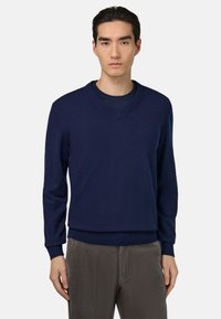 Pull à col en V bleu marine en matière douce avec manches longues et poignets côtelés, associé à un pantalon en velours côtelé gris clair.