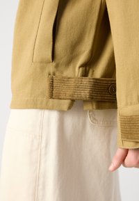 Tan cotton jacket with a corduroy accent on the belt, featuring button closure and a streamlined design. Paired with light-colored pants.