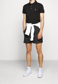 Polo noir avec un logo blanc, assorti avec un short rayé noir et blanc. Sweatshirt blanc noué autour de la taille et baskets montantes blanches.