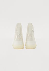 White ankle boots with a glossy finish, featuring a round logo patch, textured side perforations, and a chunky translucent sole.