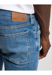 Jeans de mezclilla azul con un corte clásico, que presentan un parche de cuero marrón en la cintura trasera, cinco bolsillos y detalles de costura visibles.