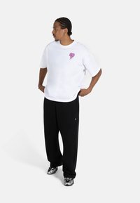 Hvit oversized t-skjorte med et rosa hjertegrafikk, paired med svarte løse bukser og sølv sneakers, som fremhever en avslappet gatekulturstil.