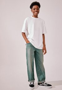 T-shirt blanche surdimensionnée, jean vert ample et baskets noires avec semelles blanches. Tenue décontractée avec une coupe relax et des manches retroussées.
