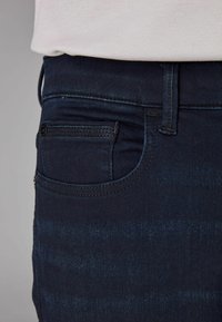 Jeans de mezclilla azul oscuro con una textura suave, que cuentan con un pequeño bolsillo frontal, pasadores de cinturón estándar y detalles de costura visibles.