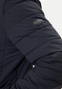 Giacca trapuntata di colore blu navy scuro con un design a quadri, texture liscia e una piccola etichetta in tessuto nero con la scritta "GUESS EST. 1981" sulla manica.