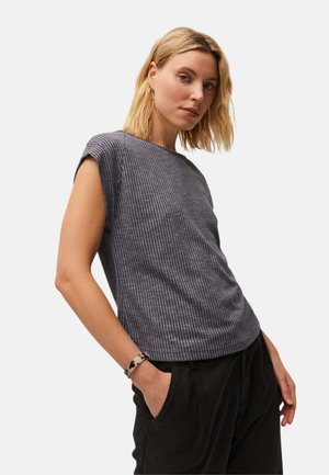 Mujer con cabello rubio que lleva una camiseta gris sin mangas con textura de canalé y pantalones negros, posando con una mano en el bolsillo frente a un fondo liso.