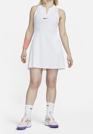 Femme portant une robe de tennis blanche sans manches de Nike, un bracelet rose, des chaussettes blanches Nike et des baskets blanches avec des accents violets et roses.