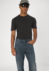 Camiseta negra de manga corta con texturizado acanalado, pantalones vaqueros de mezclilla azul con un acabado desgastado, complementados con una gorra clara y gafas de sol oscuras.