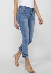Jean en denim bleu clair, coupe slim, avec un ourlet brodé présentant des motifs floraux complexes et un bord effiloché. Sandales à talons transparents.