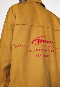 Brun tygjacka med en röd broderad design med ordet "Fusion" och flera linjer. Texturen är jämn med en hållbar yta.