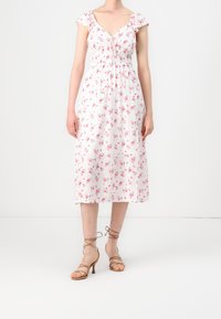 Nainen, joka käyttää valkoista midi-mekkoa, jossa on vaaleanpunaisia kukkakuoseja ja liehuvia hihoja, yhdistettynä beigeihin remmikorkokenkiin.