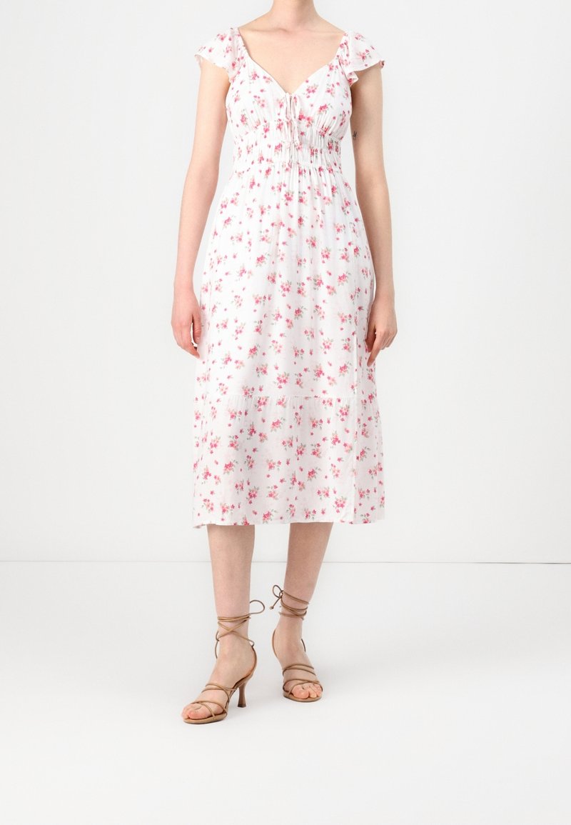 Nainen, joka käyttää valkoista midi-mekkoa, jossa on vaaleanpunaisia kukkakuoseja ja liehuvia hihoja, yhdistettynä beigeihin remmikorkokenkiin.