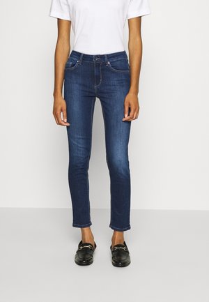 Vrouw draagt donkerblauwe skinny jeans, wit shirt met korte mouwen en zwarte instapschoenen met gouden gesp, staand tegen een witte achtergrond.
