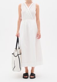 Abito bianco senza maniche con design avvolgente, vita con cordoncino e sottile rifinitura decorativa. Abbinato a una borsa a tracolla bianca con bordi neri e infradito neri.