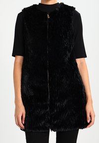 Svart faux pälsväst med hög krage och dragkedjeöppning. Har en texturerad yta och är ärmlös, kompletterad av en rak fåll.