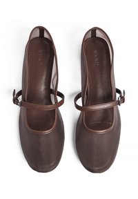Zapatos de cuero marrón con puntera redondeada, correa en el empeine y textura suave. El diseño presenta costuras expuestas y un estilo minimalista.