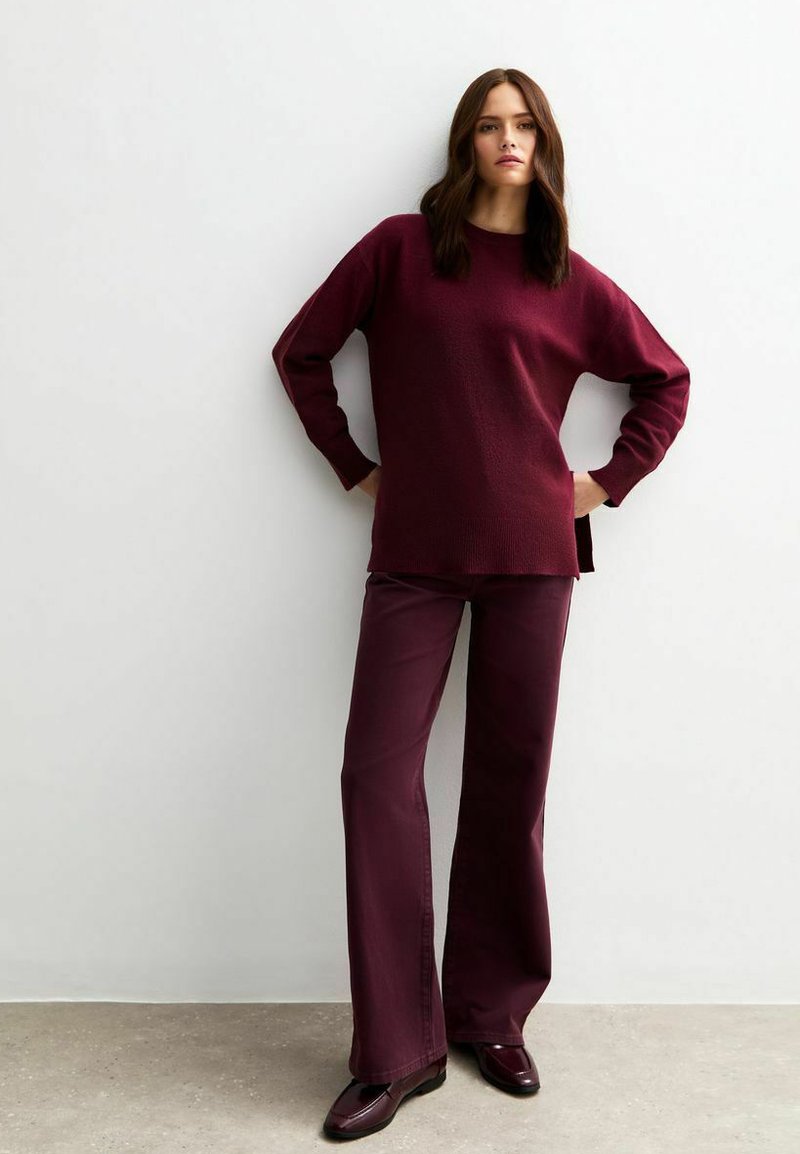 Burgundy texturerad stickad tröja med långa ärmar, tillsammans med matchande utsvängda byxor och glänsande mörk burgundy skor mot en neutral bakgrund.