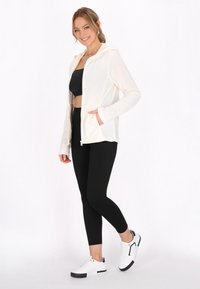 Hellbeige Zip-Up-Hoodie mit Kapuze, schwarzes enges Oberteil, schwarze Leggings und weiße Sneakers mit schwarzen Akzenten; durchgehend glatte Textur.