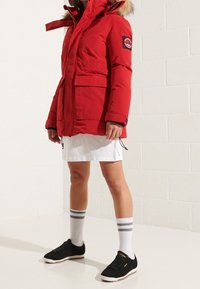 Rode gewatteerde jas met een bont gevoerde capuchon, twee voorkanten zakken en een logo patch. G gedragen met een witte rok, gestreepte sokken en zwarte sneakers.