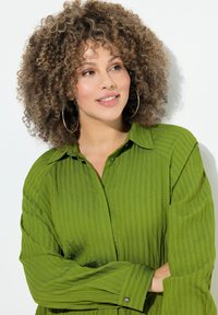 Camicia verde strutturata con colletto, maniche lunghe e dettagli con bottoni. Il modello ha i capelli ricci ed è posato su uno sfondo bianco.