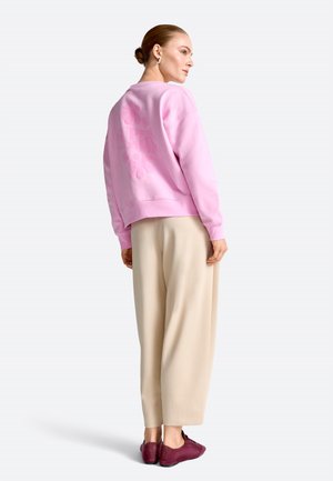 Woman with hair in bun wearing a pink sweatshirt, beige wide-leg pants, and burgundy sneakers, standing and looking sideways.