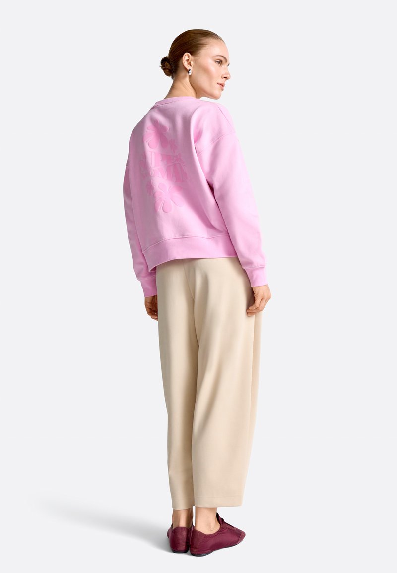 Donna con i capelli raccolti in uno chignon, che indossa una felpa rosa, pantaloni beige a gamba larga e sneakers color bordeaux, in piedi e con lo sguardo rivolto di lato.