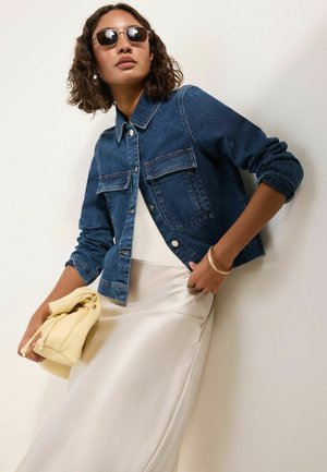 Vrouw draagt een donkerblauwe spijkerjas, witte satijnen rok, zonnebril, houdt een gele clutch tas vast, poseert tegen een lichte achtergrond.