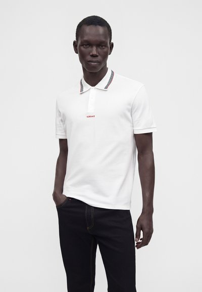 Homme portant un polo blanc Versace avec un col à motifs et un pantalon noir, debout avec une main dans la poche sur un fond uni.