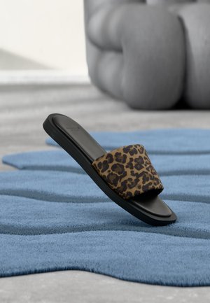 Schwarze Slidesandale mit Leopardenmuster-Riemen auf strukturiertem blauem Teppich, mit verschwommenem großen beigen Kissen im Hintergrund.